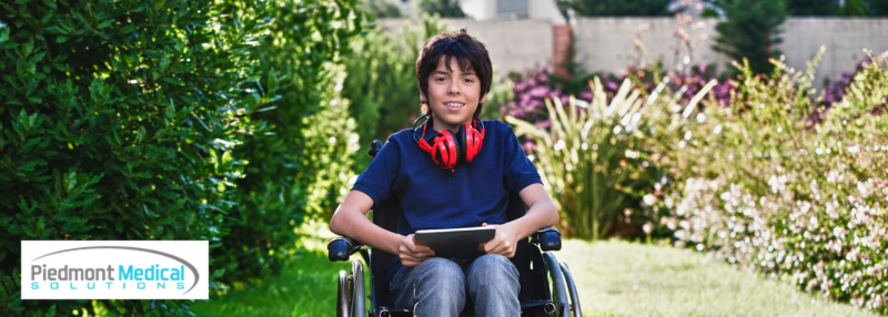 Boy in wheelchair outside smiling with headphones around his neck and tablet in his hands